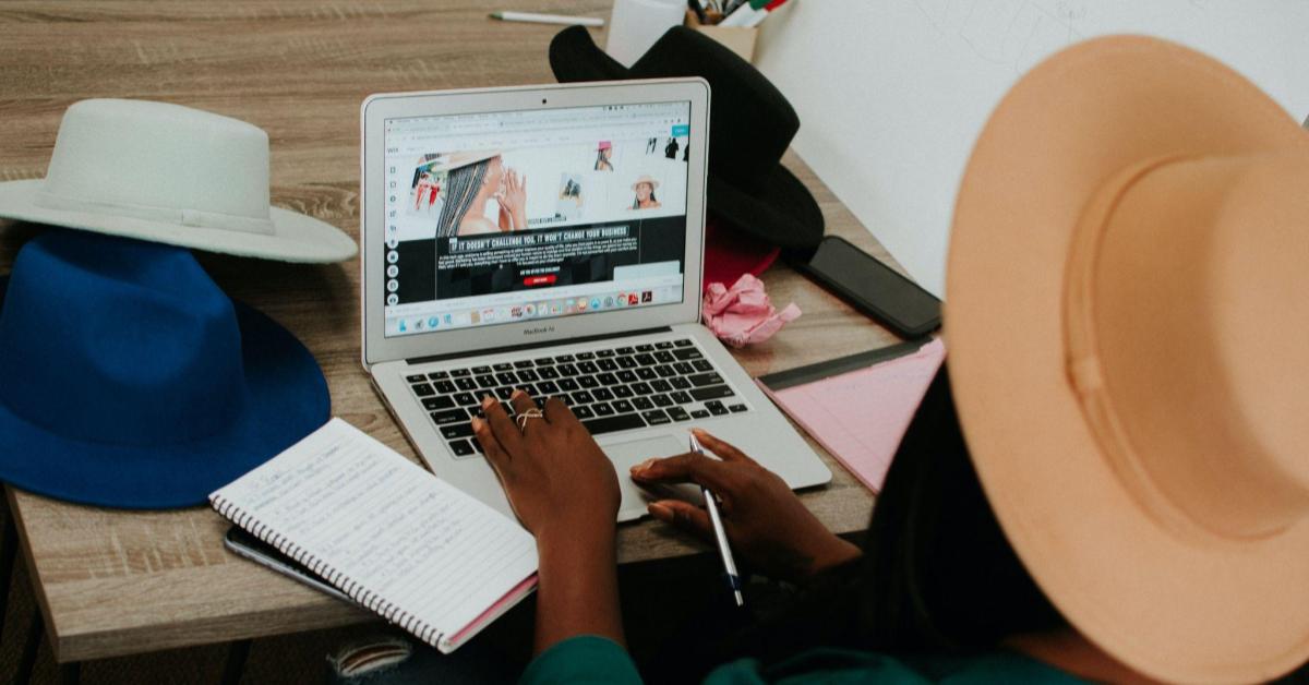 Na imagem, vemos uma mulher preta sentada numa mesa, de frente para um computador. É como se estivéssemos atrás dela, e assim, conseguimos ver o que ela observa no computador. Nele, está aberto um site de chapéus, item que a moça usa, e que também esta distribuído, em diversos modelos, na mesa. Aqui, a imagem faz alusão a um momento de estratégia de branding.